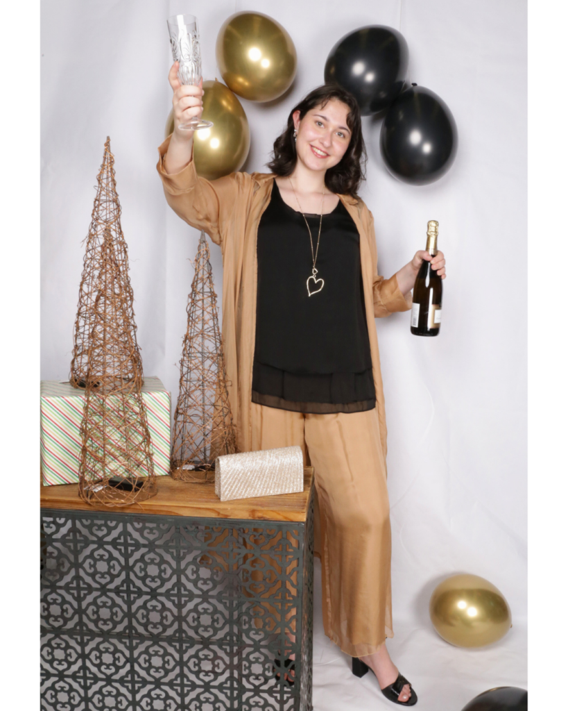 A young woman in New Year celebration outfit on a white surface surrounded by gold and black balloons, holding a champagne glass and a shimmering clutch. A bottle of champagne and decorative elements like rustic twig cones and a wrapped gift add to the celebratory atmosphere.