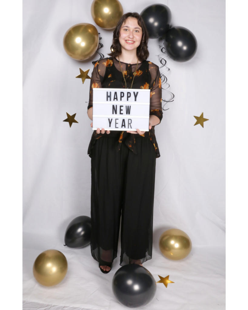 A woman in a floral-patterned outfit lies on a white sheet with black and gold balloons, golden stars, and a "HAPPY NEW YEAR" light-up sign, creating a celebratory New Year's vibe.